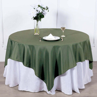 Round olive green tablecloth on a layered white table setup with flowers and glass.