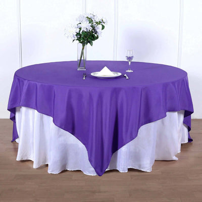 Purple round tablecloth on a white table with a vase of flowers and glass.