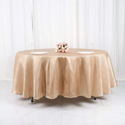 Round satin tablecloth in champagne color with floral centerpiece on a plain backdrop.