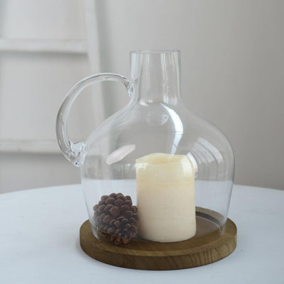 Clear glass pitcher with a candle and pinecones on a wooden base.