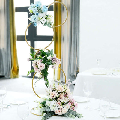 Floral centerpiece with gold rings and pastel flowers on a dining table setting.