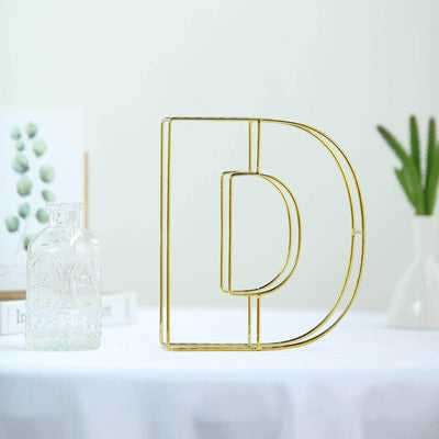 Gold letter "D" decorative piece on a table with plants in the background.