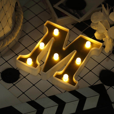 Lit marquee letter "M" with bulbs on a patterned surface.