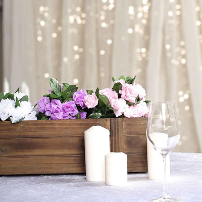 Decorative flowers in a wooden box with candles and a glass on a table.